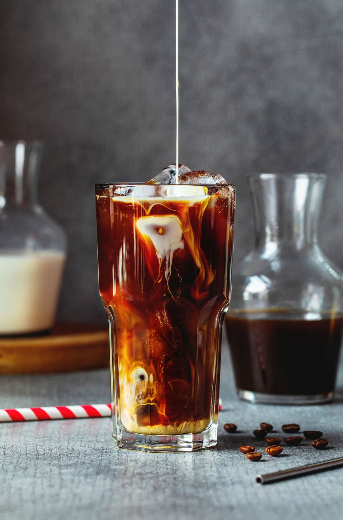 Milk poured in glass with iced coffee on table near glass jar and straw with coffee beans