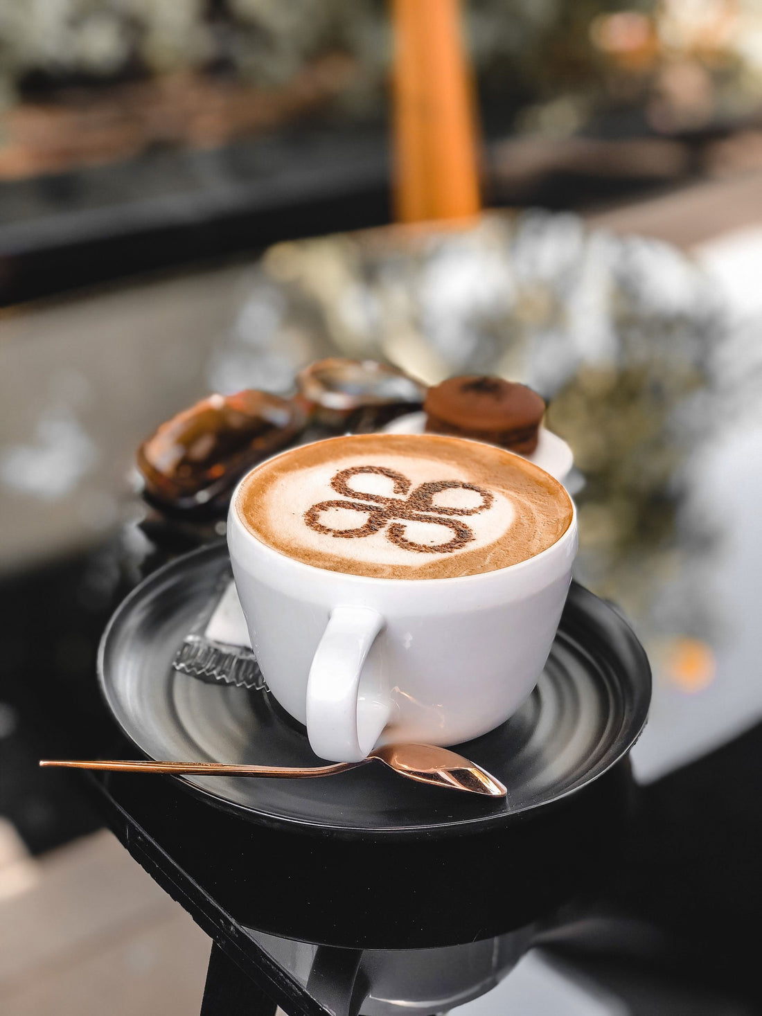 Close-up of a cappuccino with latte art on a black saucer with a teaspoon, shot outdoors.